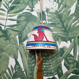 May include: White ceramic bell with a red fox design and the text "Truckee, CA". The bell has blue, green, and orange accents. It is hanging from a brown leather cord.