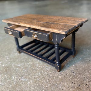 Puede incluir: Isla de cocina de madera en miniatura con dos cajones abiertos. La encimera es de color marrón claro con una textura de vetas de madera. La base y los cajones son negros con tiradores de cajón color latón. La isla tiene un estante inferior con listones.