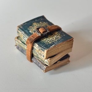 Puede incluir: Una pila de tres libros antiguos en miniatura con cubiertas verde azulado desgastadas y detalles dorados. Una correa de cuero marrón desgastada con una hebilla decorativa asegura los libros. Los bordes de las páginas están envejecidos.
