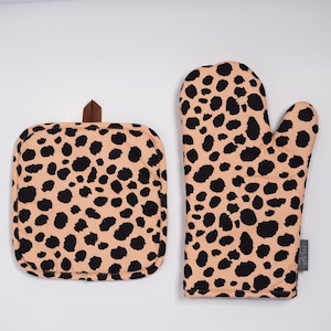 May include: A tan and black leopard print oven mitt and pot holder set. The oven mitt is a standard size, and the pot holder is square with a brown leather loop for hanging. The set is designed for kitchen use.