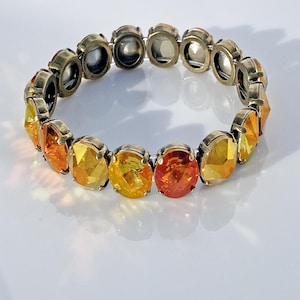 May include: A stretch bracelet featuring oval-shaped, faceted gemstones in shades of yellow and orange. The stones are set in a metal band with an antique gold finish. The bracelet is displayed on a white surface.