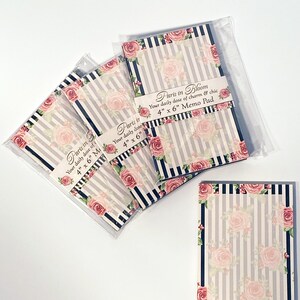 May include: Memo pads with a floral rose design and striped background. The pads are 10 cm x 15 cm and feature the text "Paris in Bloom". The pads are packaged in clear plastic.