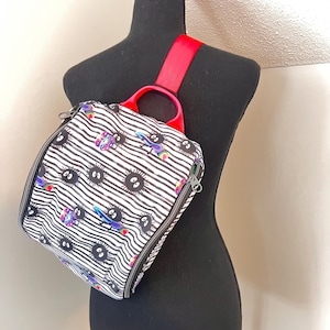 May include: A black and white striped backpack with a red strap. The backpack has a zipper closure and a handle. The backpack is decorated with a pattern of black and white eyes and colorful cartoon characters.