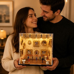 May include: A miniature art gallery diorama with gold-framed paintings, miniature figures, and a red velvet rope. The diorama is held by a couple, with the woman wearing a cream sweater and the man in a black sweater.