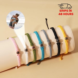 Könnte beinhalten: Sieben bunte geflochtene Armbänder mit jeweils einer dunkelgrauen Perle. Die Armbänder sind in einer Reihe auf einer weißen Oberfläche angeordnet.