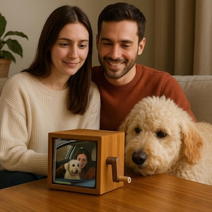 May include: A wooden photo cube with a hand crank, showing a picture of a woman and a dog in a car. The cube is on a brown table, with a couple and a fluffy dog in the background.
