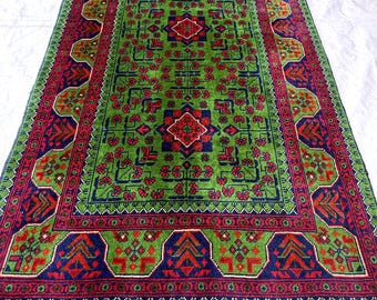 Puede incluir: Una alfombra rectangular con un campo verde vibrante y un patrón floral repetitivo en rojo y azul oscuro. La alfombra tiene un borde ancho con motivos geométricos en rojo, azul y verde, rematado con un fleco blanco corto en ambos extremos.