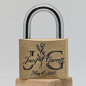 May include: A brass padlock with a silver shackle. The lock is engraved with the names "Jacy" and "Garrett", a floral design, and the date "May 6th, 2017". The lock is square and sits on a wooden block.