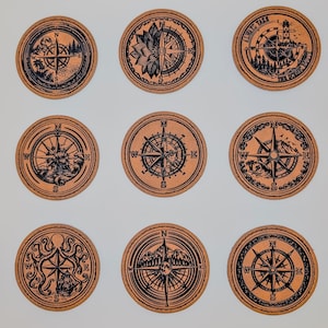 May include: Nine round coasters with compass designs. Each coaster features a different black compass rose illustration on a brown background. Some designs include nautical elements like ships, lighthouses, and mountains. The coasters are arranged in a grid.