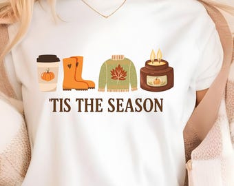 May include: White t-shirt with fall-themed graphics, including a coffee cup with a pumpkin, orange rain boots, a green sweater with a leaf, and a candle. The text "'Tis the Season" is printed below the graphics.
