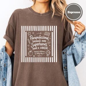 Paraprofessional Comfort Colors espresso shirt featuring the phrase "Paraprofessional Because Even Superheroes Need a Sidekick" with school-themed doodles including books, pencil, apple, globe, hearts, and stars in a hand-drawn style.
