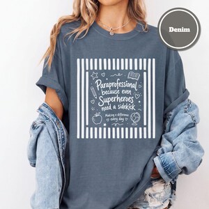 以下が含まれることがあります： ストライプの縁取りが施された白いグラフィックプリントのデニムカラーのTシャツ。テキストは「Paraprofessional because even Superheroes need a sidekick, Making a difference every day」と書かれており、鉛筆、本、リンゴ、地球儀のアイコンが添えられています。