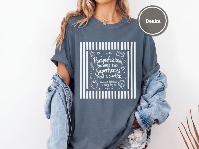 以下が含まれることがあります： ストライプの縁取りが施された白いグラフィックプリントのデニムカラーのTシャツ。テキストは「Paraprofessional because even Superheroes need a sidekick, Making a difference every day」と書かれており、鉛筆、本、リンゴ、地球儀のアイコンが添えられています。