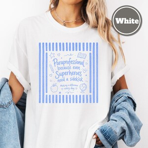以下が含まれることがあります： 両サイドに縦縞模様が入った青いグラフィックプリントの白いTシャツ。テキストには「Paraprofessional because even Superheroes need a sidekick, Making a difference every day」と書かれており、鉛筆、本、リンゴ、地球儀のアイコンが添えられています。
