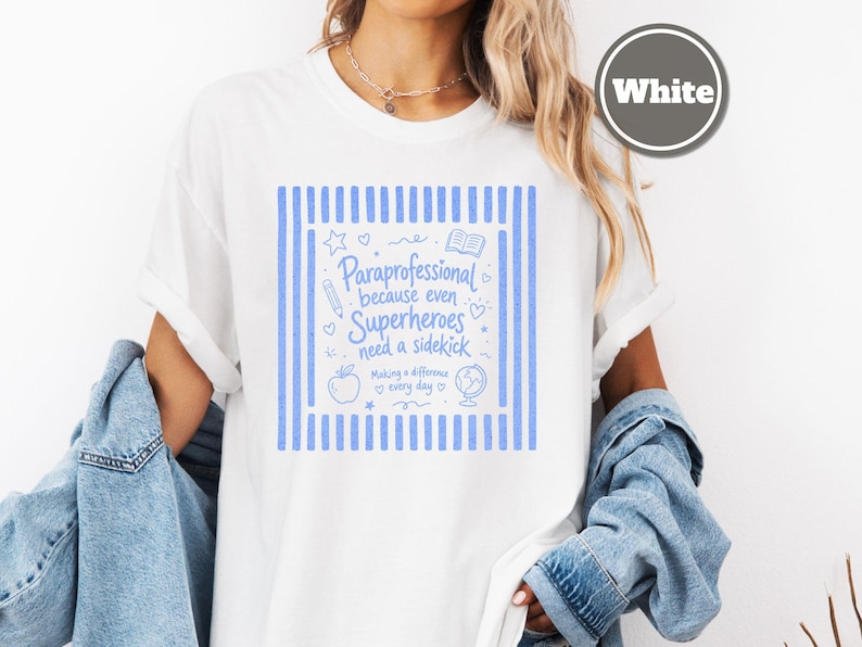 以下が含まれることがあります： 両サイドに縦縞模様が入った青いグラフィックプリントの白いTシャツ。テキストには「Paraprofessional because even Superheroes need a sidekick, Making a difference every day」と書かれており、鉛筆、本、リンゴ、地球儀のアイコンが添えられています。