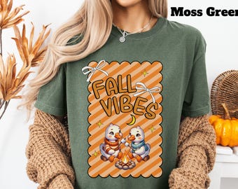 May include: Moss green t-shirt with a Fall Vibes graphic. The graphic features two cartoon ducks roasting marshmallows over a campfire, with the words "Fall Vibes" in orange letters. The design is set against an orange and white striped background.