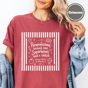 以下が含まれることがあります： クリムゾン色の半袖Tシャツ。垂直のストライプが入った白いグラフィックプリントが施されており、「Paraprofessional because even Superheroes need a sidekick, Making a difference every day」というテキストと、鉛筆、本、リンゴ、地球儀のアイコンが描かれています。