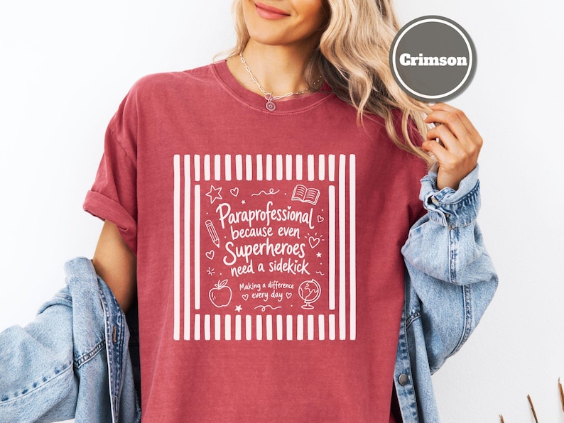 以下が含まれることがあります： クリムゾン色の半袖Tシャツ。垂直のストライプが入った白いグラフィックプリントが施されており、「Paraprofessional because even Superheroes need a sidekick, Making a difference every day」というテキストと、鉛筆、本、リンゴ、地球儀のアイコンが描かれています。
