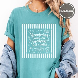 以下が含まれることがあります： シーフォームグリーンのTシャツで、両サイドに縦縞模様の白いグラフィックプリントが施されています。テキストは「Paraprofessional because even Superheroes need a sidekick, Making a difference every day」と書かれており、鉛筆、本、リンゴ、地球儀のアイコンが添えられています。