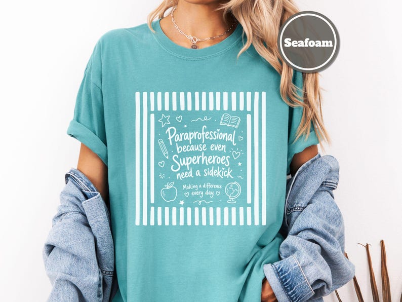 以下が含まれることがあります： シーフォームグリーンのTシャツで、両サイドに縦縞模様の白いグラフィックプリントが施されています。テキストは「Paraprofessional because even Superheroes need a sidekick, Making a difference every day」と書かれており、鉛筆、本、リンゴ、地球儀のアイコンが添えられています。