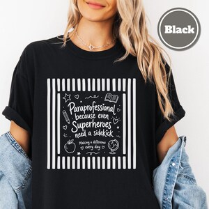 以下が含まれることがあります： 両サイドに縦縞が入った白いグラフィックデザインの黒いTシャツ。テキストは「Paraprofessional because even Superheroes need a sidekick. Making a difference every day.」と書かれています。鉛筆、本、リンゴ、地球儀のアイコンが含まれています。