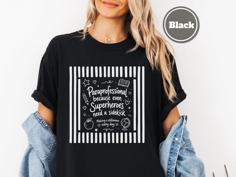 以下が含まれることがあります： 両サイドに縦縞が入った白いグラフィックデザインの黒いTシャツ。テキストは「Paraprofessional because even Superheroes need a sidekick. Making a difference every day.」と書かれています。鉛筆、本、リンゴ、地球儀のアイコンが含まれています。