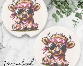 May include: Two white ceramic coasters with a pink and brown cartoon Highland cow wearing sunglasses and a pink bandana. The cow is holding a football and the text "Sierra" is written below the cow. The coasters are personalized.