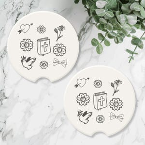 May include: Two off-white car coasters with black line art. Each coaster features a heart with an arrow, flowers, a book with a cross, a dove, and a bow. The coasters are round with a small notch for easy removal.