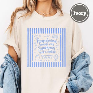 以下が含まれることがあります： アイボリーのTシャツで、縦縞の青いグラフィックプリントと「Paraprofessional because even Superheroes need a sidekick」というテキスト、さらに鉛筆、本、リンゴ、地球儀、星、ハートの小さなイラストが描かれています。