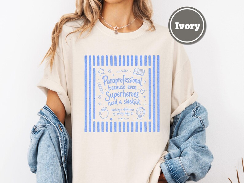 以下が含まれることがあります： アイボリーのTシャツで、縦縞の青いグラフィックプリントと「Paraprofessional because even Superheroes need a sidekick」というテキスト、さらに鉛筆、本、リンゴ、地球儀、星、ハートの小さなイラストが描かれています。