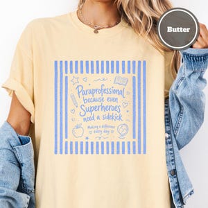 以下が含まれることがあります： バターイエローのTシャツ。青いグラフィックプリントには「Paraprofessional because even Superheroes need a sidekick」および「Making a difference every day」という文字が記され、鉛筆、リンゴ、地球儀、本のアイコンが周囲に配置されています。
