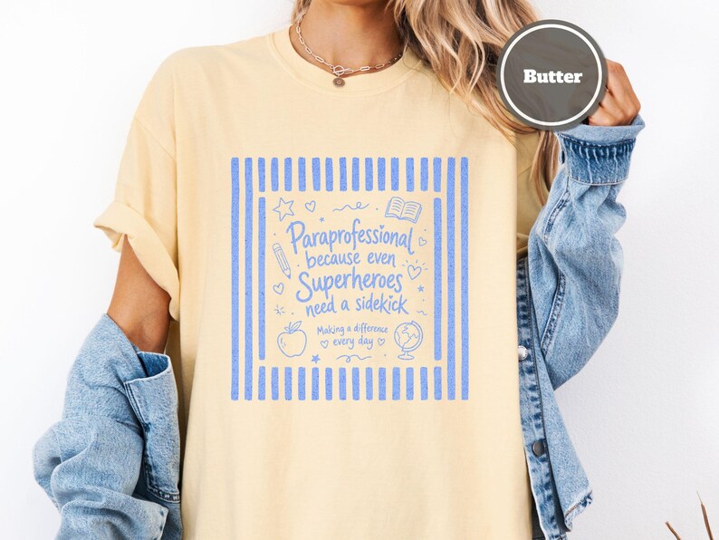 以下が含まれることがあります： バターイエローのTシャツ。青いグラフィックプリントには「Paraprofessional because even Superheroes need a sidekick」および「Making a difference every day」という文字が記され、鉛筆、リンゴ、地球儀、本のアイコンが周囲に配置されています。