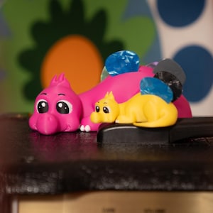 Pode incluir: Um conjunto de figuras de dinossauros rosa e amarelo. O dinossauro maior é rosa com um acento azul claro e o dinossauro menor é amarelo. Os dinossauros estão sentados em uma superfície preta.