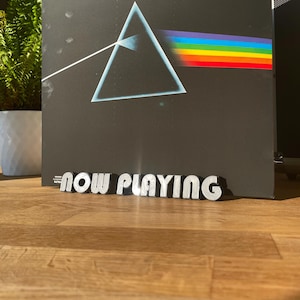 Op de afbeelding: Een zwarte vinylplaat met een prisma- en driehoeksontwerp. De plaat is getiteld "Dark Side of the Moon" van Pink Floyd. Voor de plaat staat een zilveren bord met de tekst "NOW PLAYING".