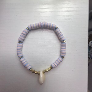 May include: A beaded bracelet featuring light blue and white striped beads, interspersed with gold-colored beads. A white teardrop-shaped pendant hangs from the center. The bracelet is displayed on a white surface.