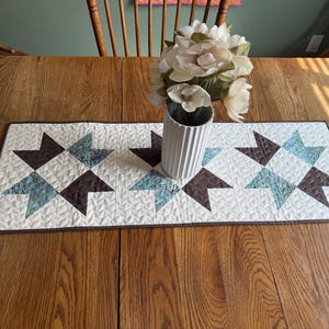 May include: A rectangular quilted table runner featuring three star-shaped blocks in shades of dark brown and teal blue on a cream background. The runner has a dark brown border and is decorated with a white ribbed vase containing white flowers.