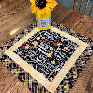 May include: A black and yellow plaid table runner with a black and white fabric panel featuring a Thanksgiving theme. The panel has a chalkboard design with the words "happy thanksgiving", "grateful", "blessed", and "give thanks".