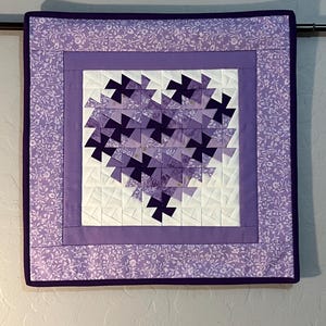May include: A square quilted wall hanging with a heart design. The heart is made of dark purple and light purple fabric pieces. The heart is centered on a white background, framed by a purple border and a floral patterned border.