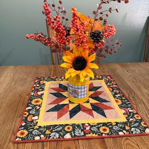 May include: A yellow metal container with the text Hello Fall and a truck graphic, holding a bouquet of artificial sunflowers, orange berries, and pinecones. It sits on a square quilted fabric mat featuring a star pattern and mushroom print.