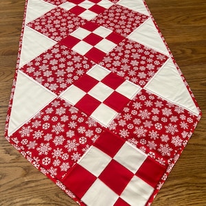 May include: A red and white quilted table runner with a festive design. The runner features a pattern of red squares, white squares, and red fabric with white snowflakes. The edges are finished with a red border, creating a seasonal accent for any table.