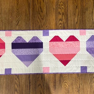 May include: A quilted table runner featuring four heart shapes in shades of pink and purple. The hearts are made of fabric strips and set against a white background with a scalloped edge. The runner is approximately 12 inches wide.