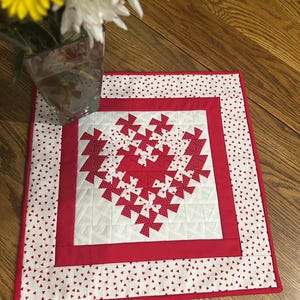 May include: A square, handmade quilt with a red and white heart design. The heart is centered on a white background, surrounded by a red border and a white border with red triangle accents. The quilt is likely used as a table runner or placemat.