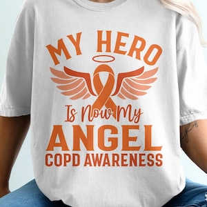 May include: White t-shirt featuring the orange text "MY HERO Is Now My ANGEL COPD AWARENESS". The design includes an orange ribbon with angel wings and a halo. The shirt is a casual style.