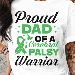 Op de afbeelding: Wit T-shirt met zwarte tekst die "Proud Dad of a Cerebral Palsy Warrior" luidt. De tekst is versierd met groene sterren en een groen lint met een verenpatroon.
