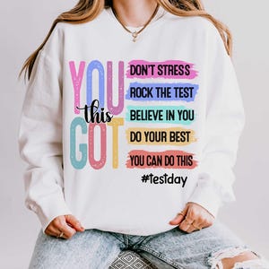 Puede incluir: Una sudadera blanca de manga larga con el texto grande y colorido YOU GOT y el texto más pequeño en negro this. A la derecha, cinco pinceladas horizontales en rosa, azul, turquesa, amarillo y rojo contienen frases motivadoras y #testday.