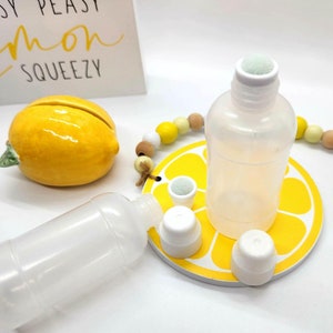 Pode incluir: Duas garrafas de plástico transparente com tampas brancas e um aplicador de esponja. As garrafas estão sobre um apoio de copo amarelo em forma de limão com detalhes brancos. As garrafas estão vazias e prontas para serem preenchidas com líquido.