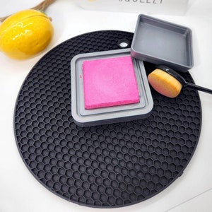 May include: A pink ink pad in a grey plastic case with a black silicone mat and a brush. The ink pad is on a black silicone mat with a honeycomb pattern. The text "Easy Peasy Lemon Squeezy" is on a white sign.