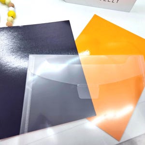 May include: A collection of stationery items, including a dark blue glossy paper, an orange paper, and a clear plastic envelope. The items are arranged on a white surface, with a wooden bead garland in the upper left corner.