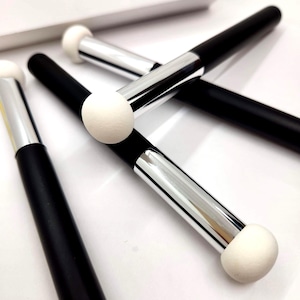 May include: Close-up of several black and silver makeup applicators with white sponge tips. The applicators are arranged diagonally across a white surface. The word "SQUEEZY" is partially visible in the background.