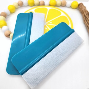 Op de afbeelding: Twee turquoise rakelen met witte vilten randen worden getoond. Ze liggen op een wit oppervlak, met een citroenontwerp en het woord "Squeezy" op de achtergrond. Een kralenketting met gele en houten kralen is ook zichtbaar.